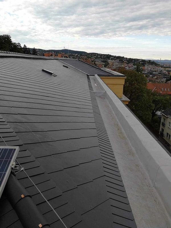 Sötétszürke, kiselemes tetőfedés közelről, a háttérben a város